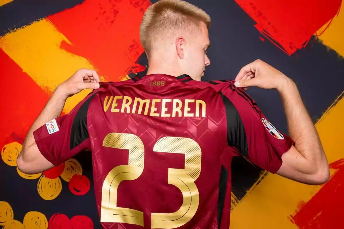 Un jugador de fútbol muestra la parte trasera de su camiseta, que tiene el nombre "Vermeeren" y el número 23, con un fondo colorido.