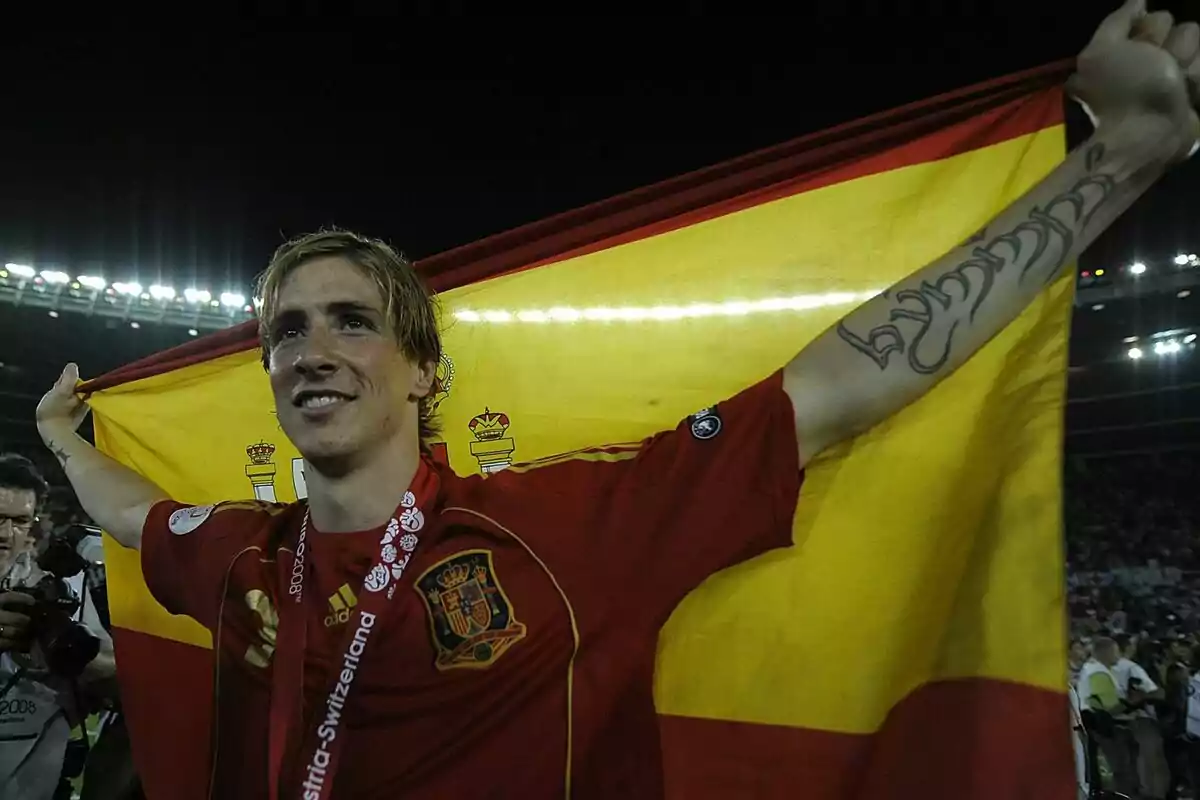 Un jugador de fútbol con la camiseta de la selección española sostiene una bandera de España en un estadio iluminado.