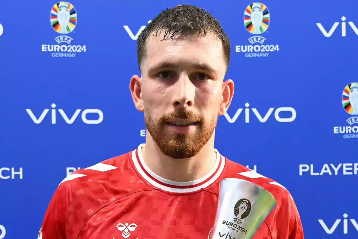 Un jugador de fútbol con uniforme rojo sostiene un trofeo frente a un fondo azul con logotipos de la UEFA Euro 2024 y Vivo.