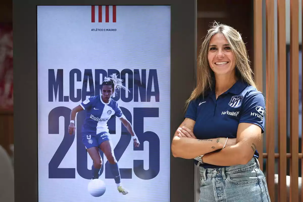 Una jugadora de fútbol sonríe junto a un cartel que muestra su imagen y el texto 